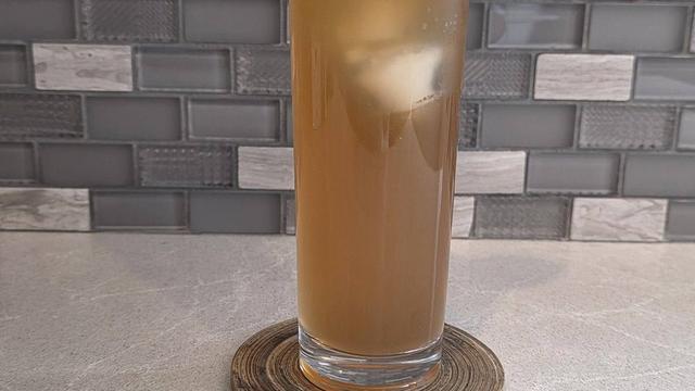 Bartesian 19th Hole cocktail, a golden brown lemonade and iced tea drink over ice in a tall highball glass with a lemon slice on the rim