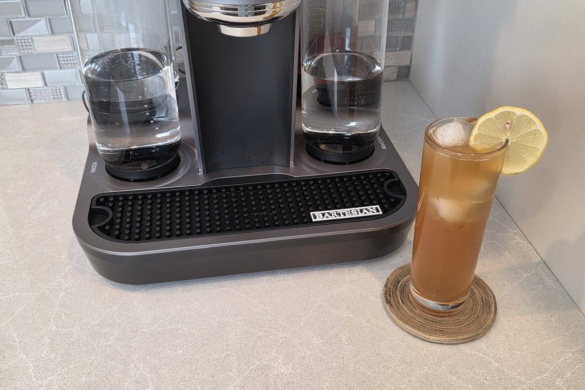 The finished 19th Hole cocktail in a tall highball glass with a lemon slice, standing beside the Bartesian machine on a home bar
