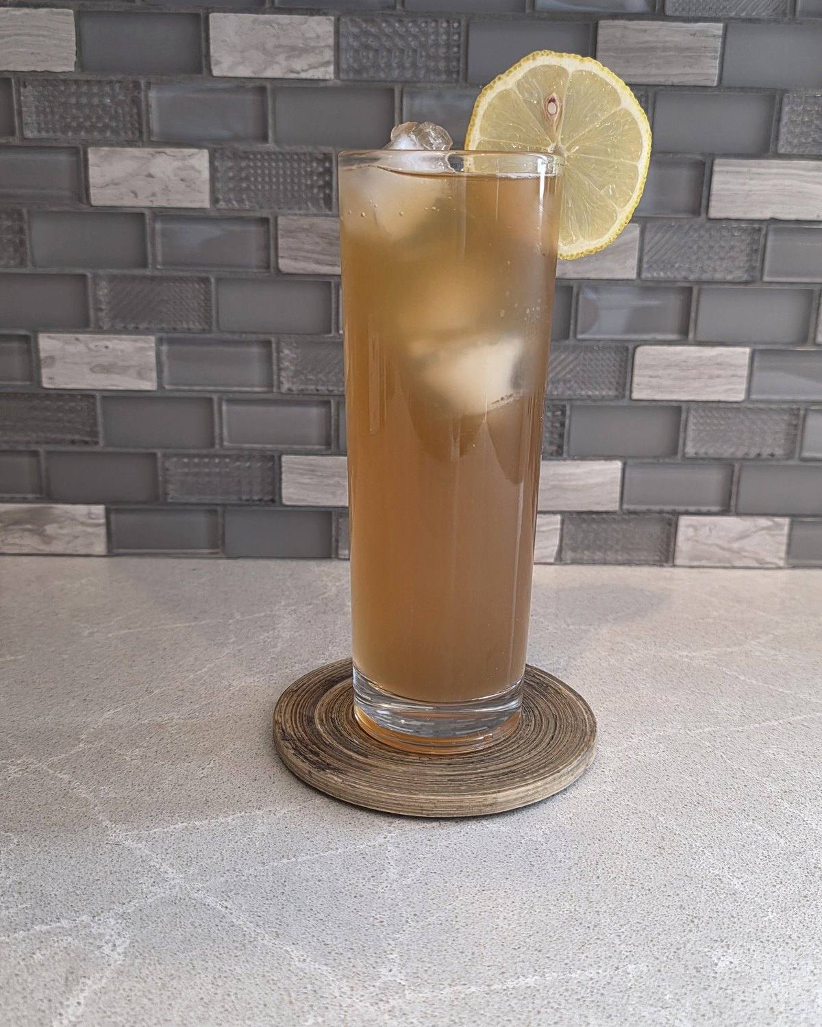 Bartesian 19th Hole cocktail, a golden brown lemonade and iced tea drink over ice in a tall highball glass with a lemon slice on the rim