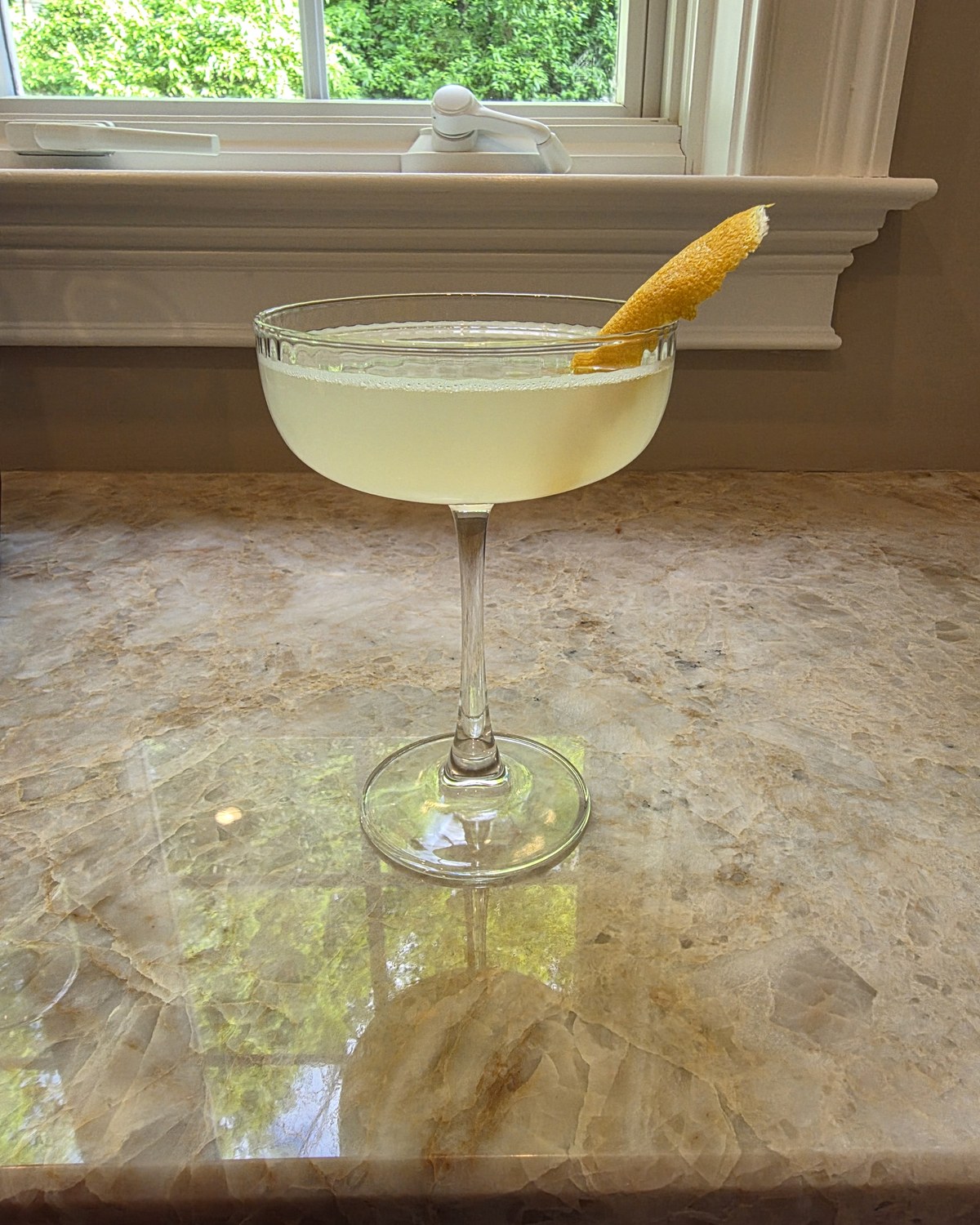Bartesian Bee's Knees cocktail in a coupe glass with a pale yellow color and a lemon peel garnish, served on a travertine counter by a sunny window