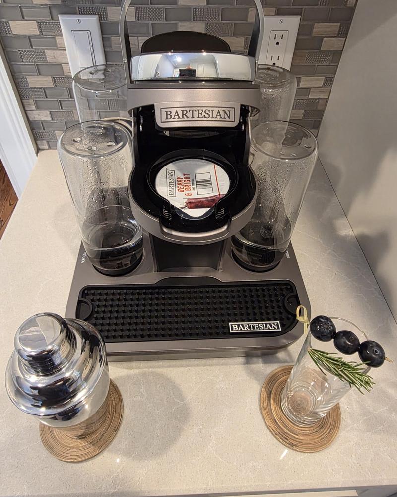 A Bartesian Premier with its lid open and the Berry & Bright capsule loaded, with a cocktail shaker and an empty highball glass holding a blueberry and rosemary pick in front