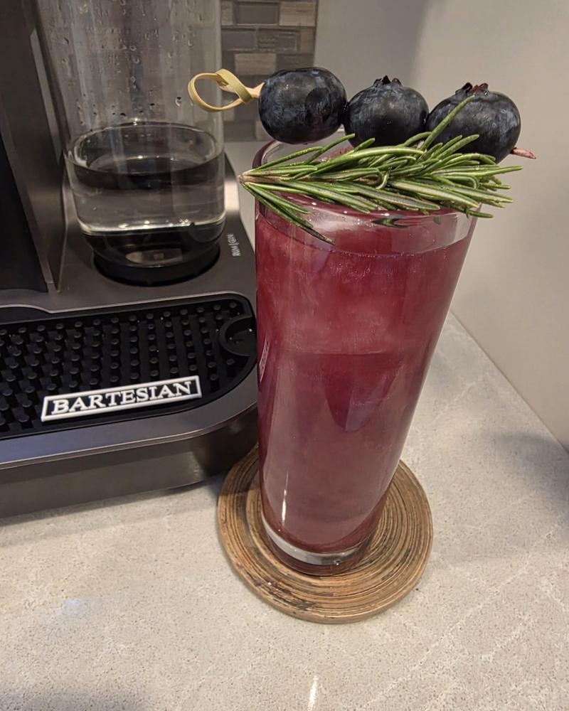 A Berry & Bright cocktail in a tall highball glass on a wooden coaster beside a Bartesian machine, with three blueberries on a pick and a rosemary sprig across the rim