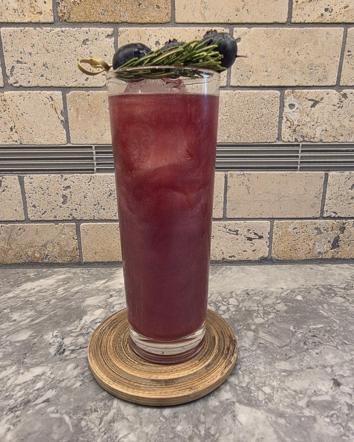 A deep cranberry Berry & Bright cocktail over ice in a tall highball glass on a wooden coaster, with three blueberries on a pick and a rosemary sprig laid across the rim
