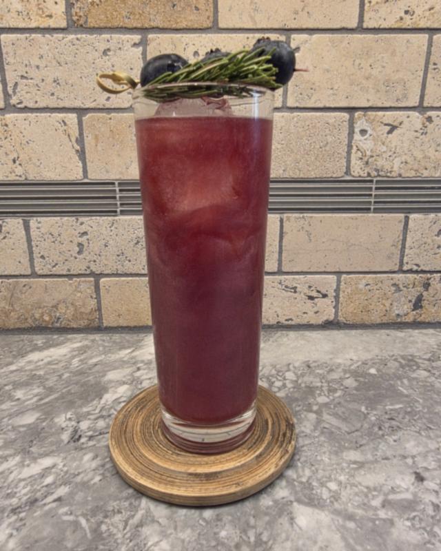 A deep cranberry Berry & Bright cocktail over ice in a tall highball glass on a wooden coaster, with three blueberries on a pick and a rosemary sprig laid across the rim