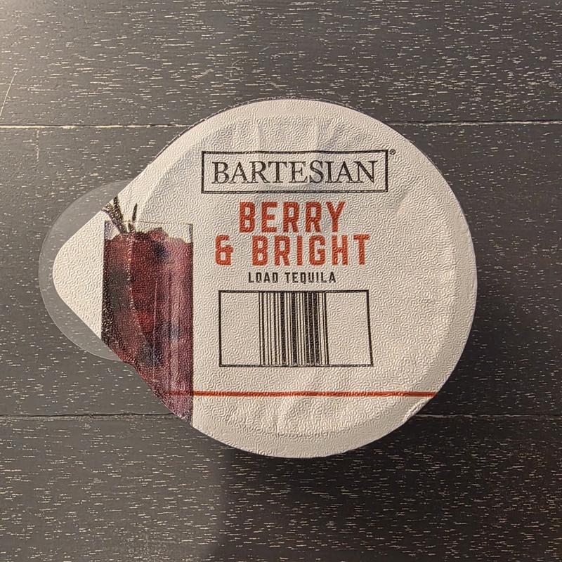 The Bartesian Berry & Bright capsule lid on a dark wood table, with the Load Tequila instruction, a drawing of the drink and the barcode visible