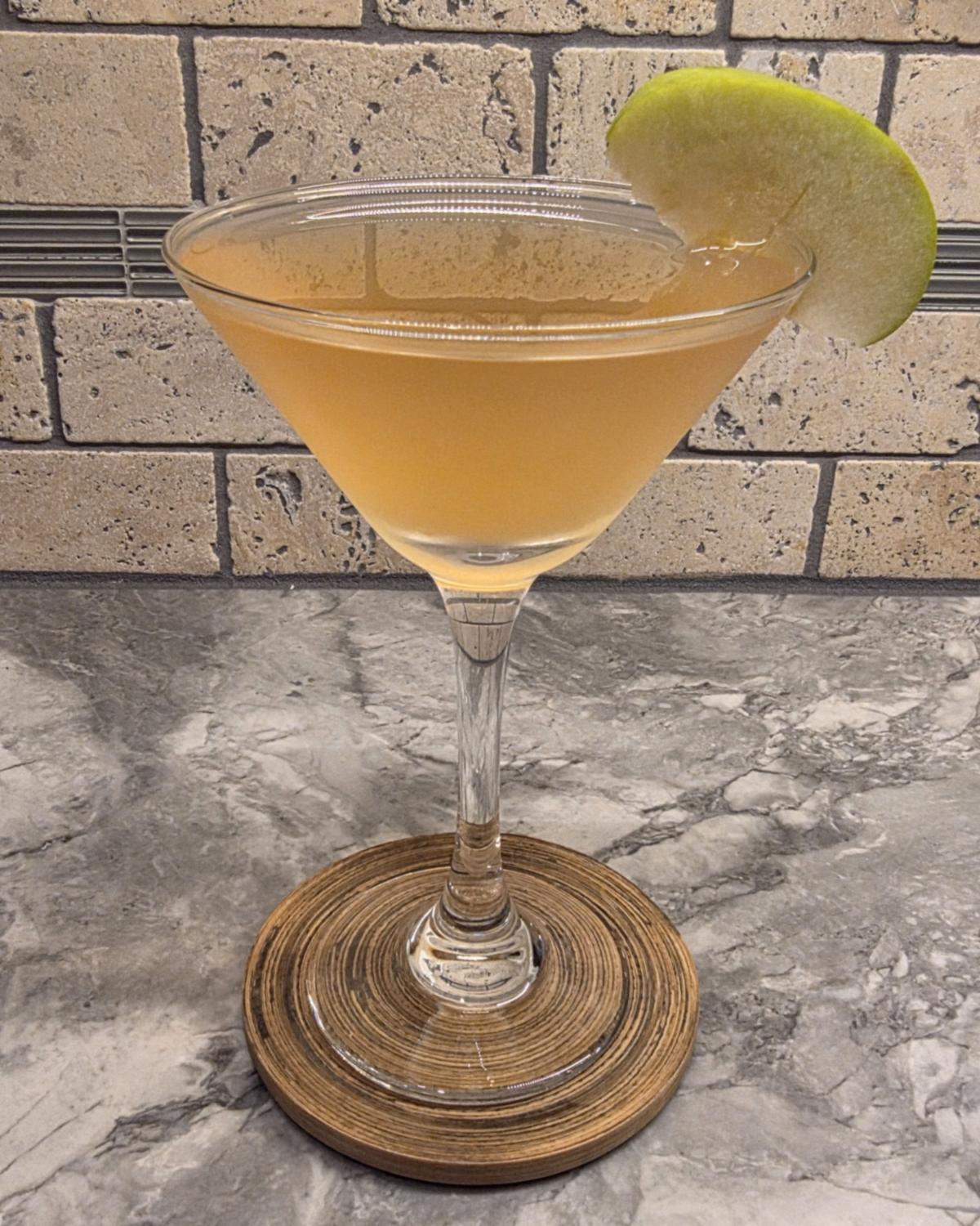 A Caramel Appletini in a stemmed martini glass on a wooden coaster, with a green apple slice on the rim