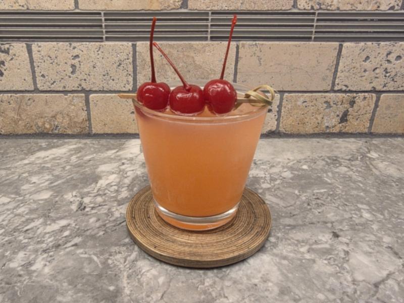 A Frosted Ambrosia in a lowball glass on a woven coaster, with three maraschino cherries on a pick, on a gray stone counter