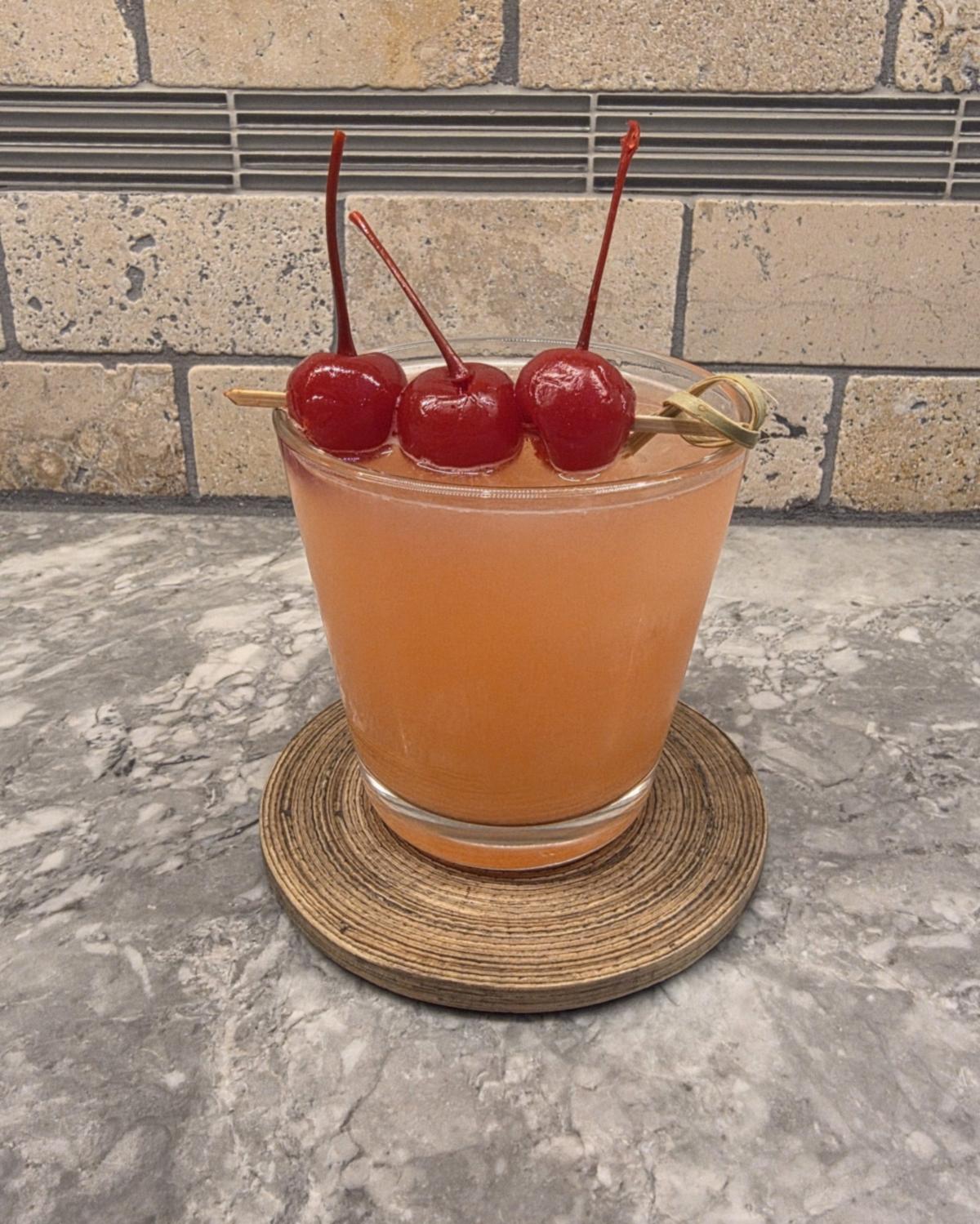 A light orange Frosted Ambrosia cocktail in a lowball glass on a woven coaster, with three maraschino cherries on a pick across the rim