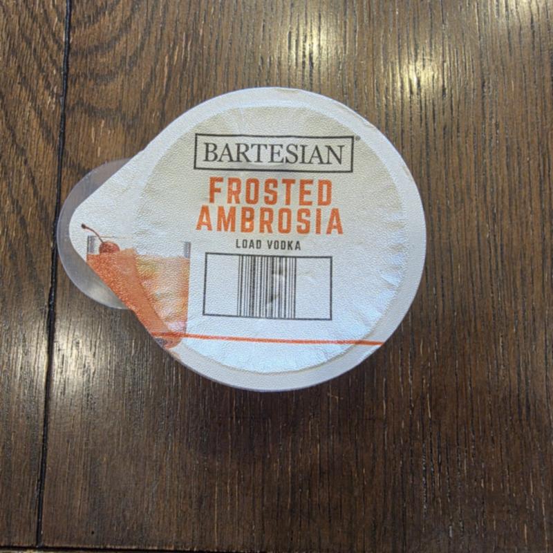 The Bartesian Frosted Ambrosia capsule lid on a dark wood table, with the Load Vodka instruction and barcode visible