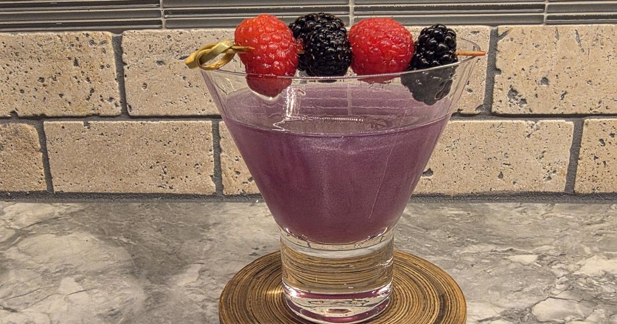 A purple, shimmering Witch's Heart cocktail in a stemless martini glass with a pick of raspberries and blackberries across the rim