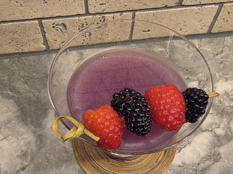 Looking down into a Witch's Heart cocktail, showing the purple shimmer swirling under a pick of raspberries and blackberries