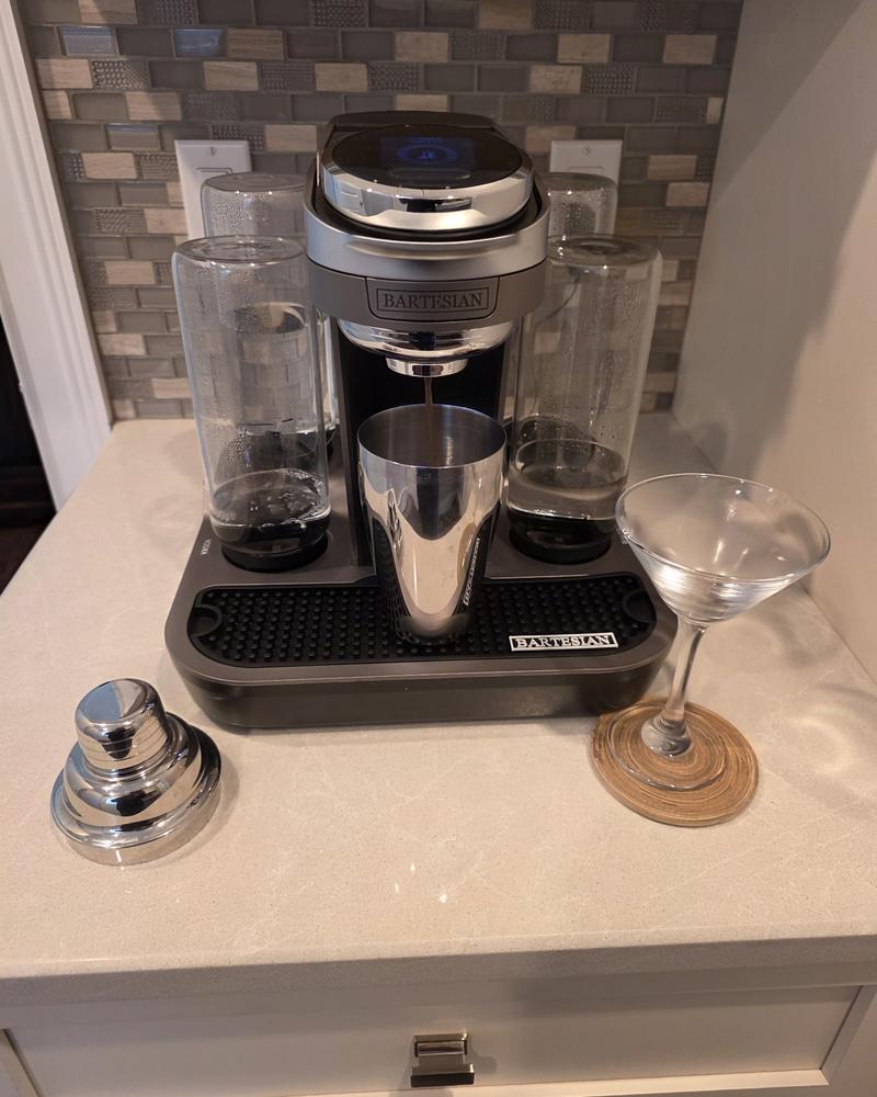 A Bartesian Premier with a stainless steel shaker under the spout, beside an empty martini glass on a woven coaster