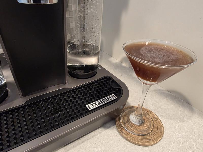 A Salted Caramel Espresso Martini in a martini glass on a woven coaster, beside the drip tray of a Bartesian machine