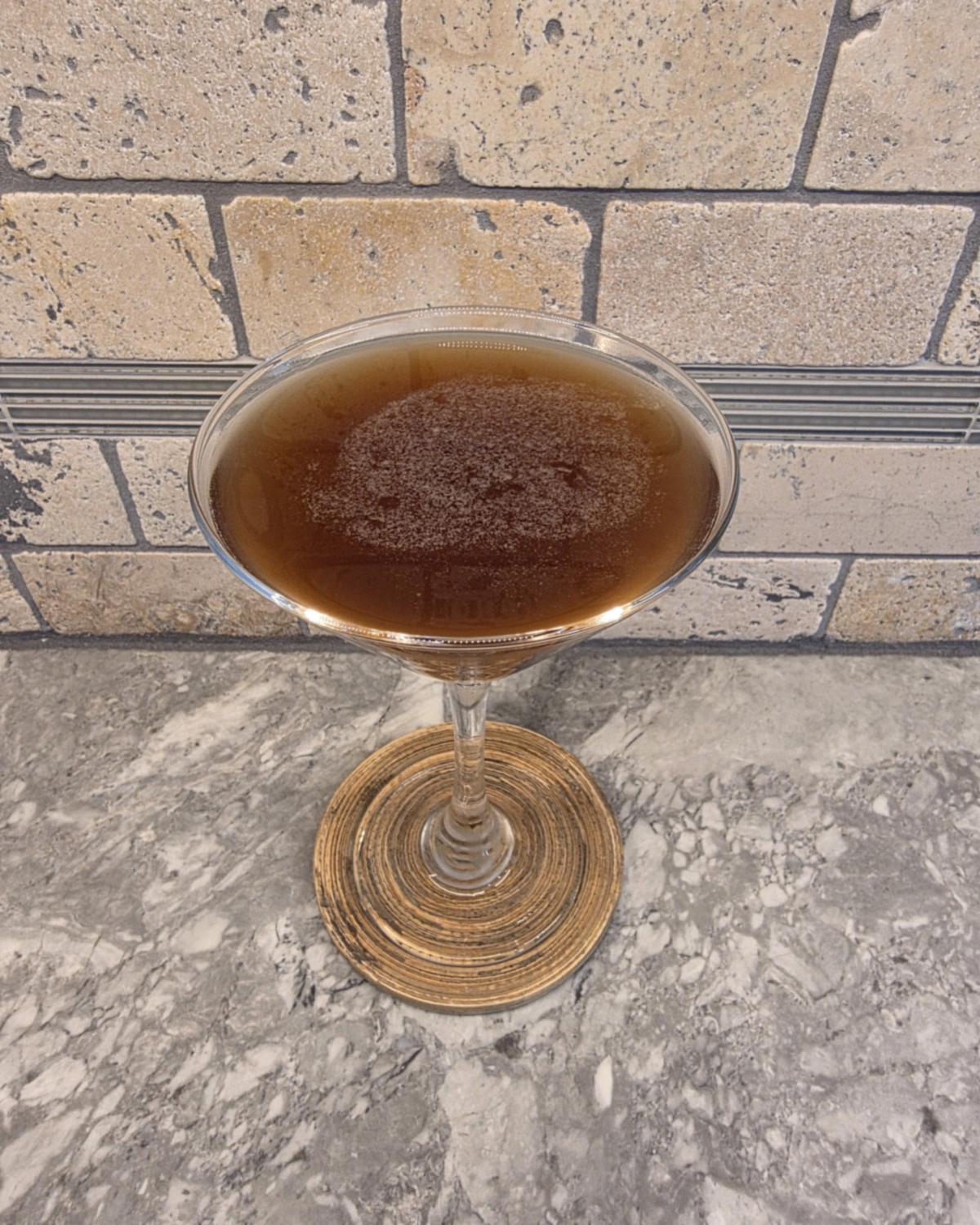 A Salted Caramel Espresso Martini in a martini glass on a woven coaster, deep brown with a little foam on top, in front of a stone tile wall