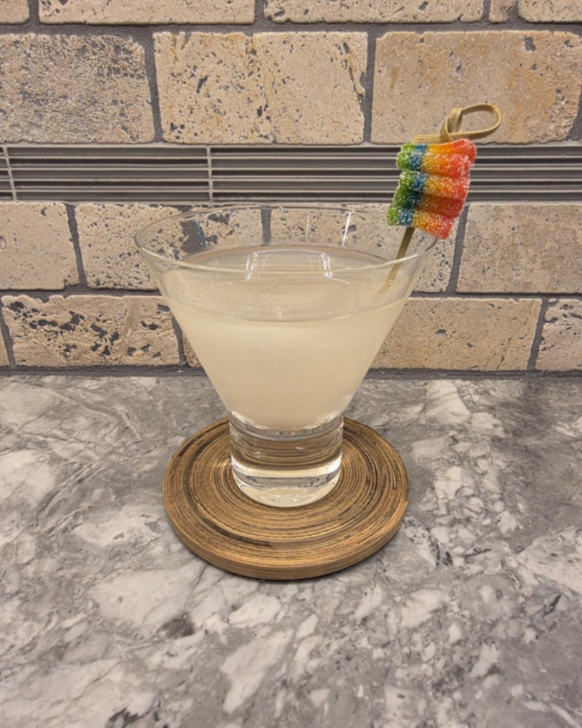 A pale, shimmery Sour Spell cocktail in a stemless martini glass on a wooden coaster, with rainbow sour candy on a pick at the rim