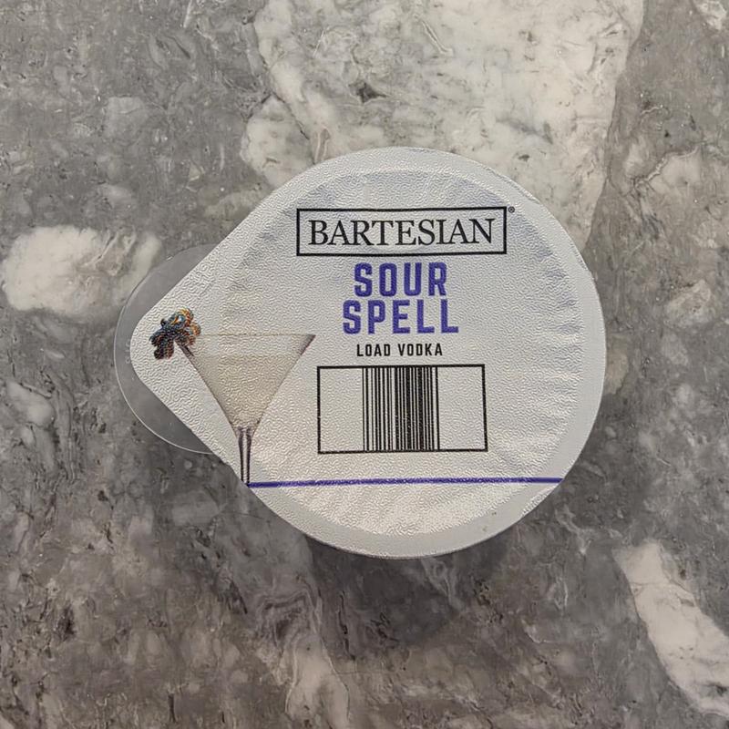 The Bartesian Sour Spell capsule lid on a marble counter, with the Load Vodka instruction and barcode visible