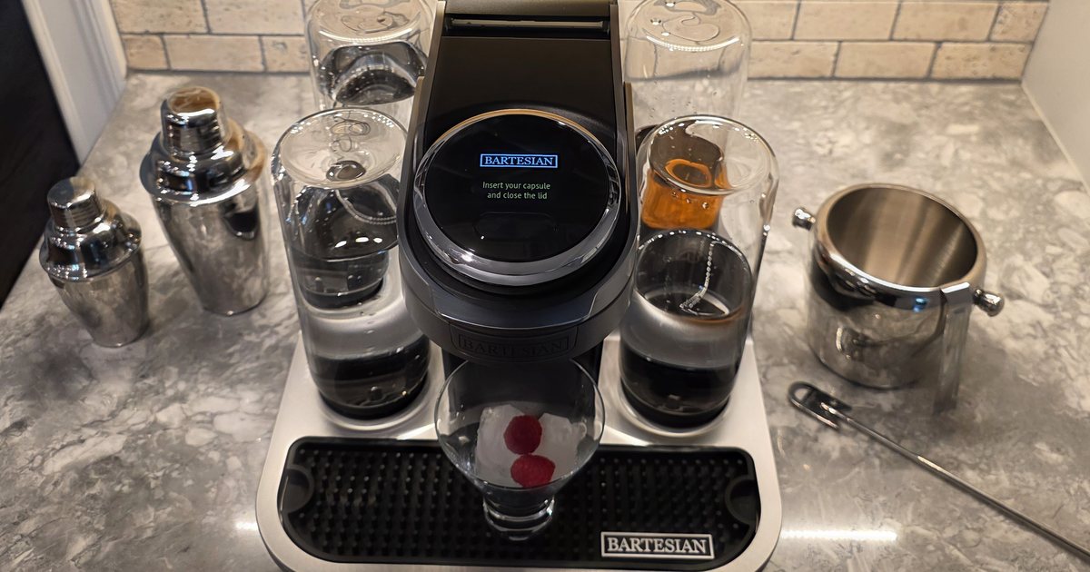 A Bartesian cocktail maker on a marble counter, with two cocktail shakers, glassware, an ice bucket and a bar spoon set out beside it.