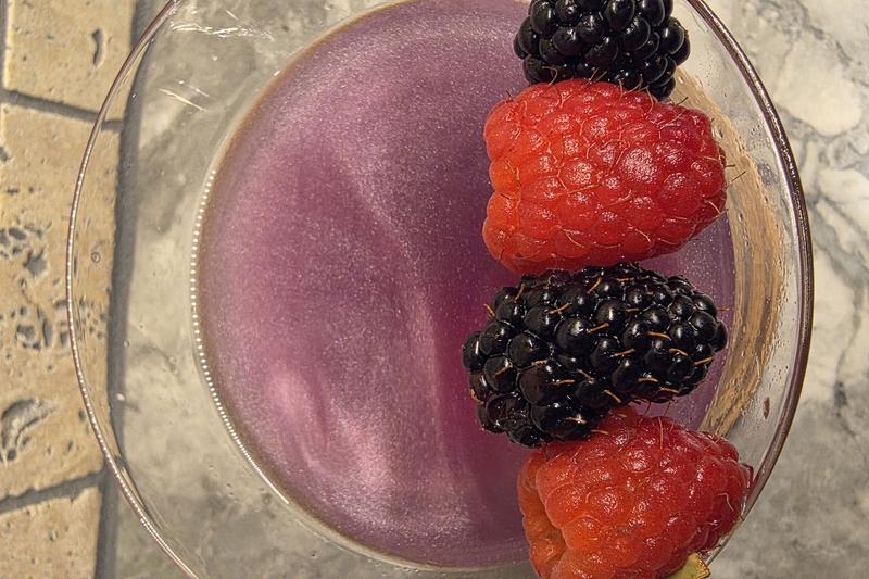 Looking down into a Witch's Heart cocktail, the purple shimmer visible beneath raspberries and blackberries on a pick