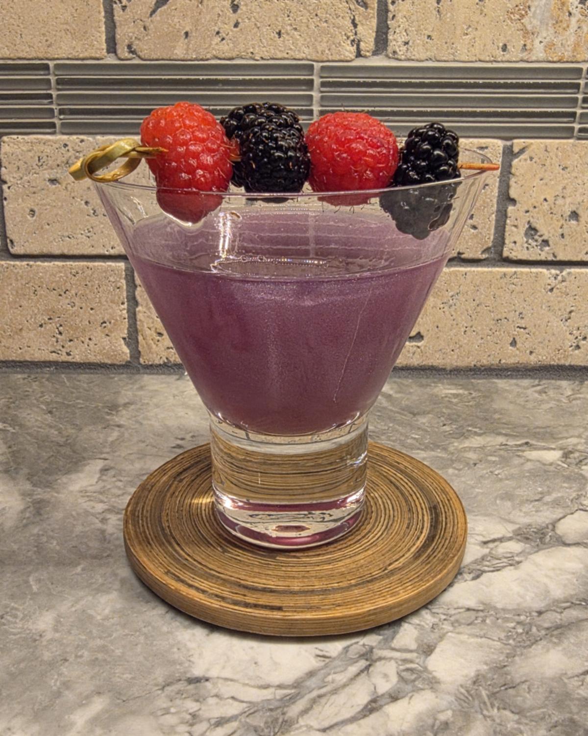 A purple, shimmering Witch's Heart cocktail in a stemless martini glass on a wooden coaster, with raspberries and blackberries on a pick across the rim