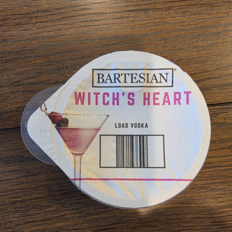 The Bartesian Witch's Heart capsule lid on a wooden table, with the Load Vodka instruction and barcode visible