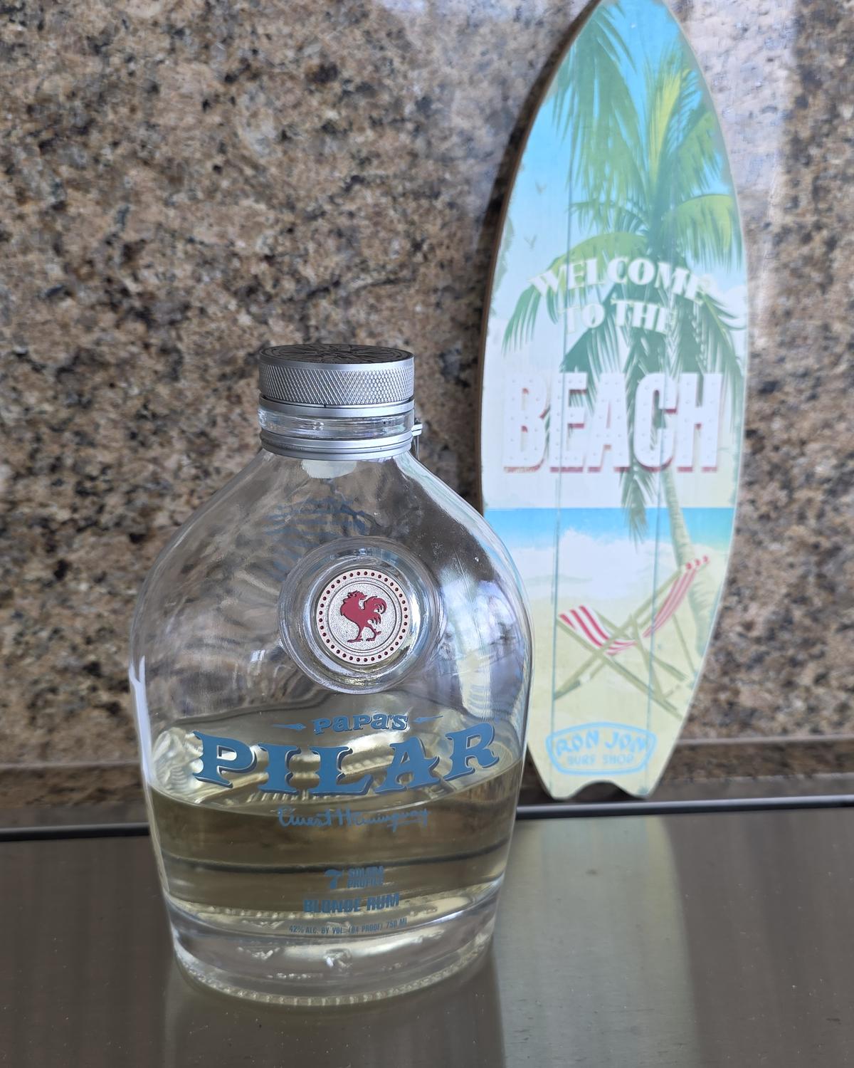 A canteen-shaped bottle of Papa's Pilar Blonde rum in front of a small Welcome to the Beach surfboard sign