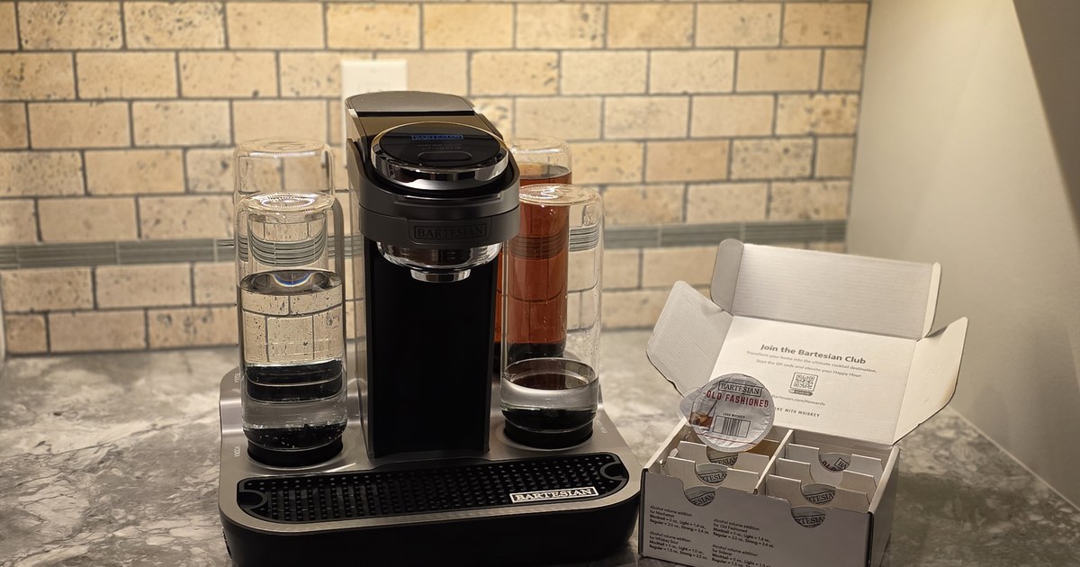 Bartesian cocktail machine on a granite countertop next to an open Bartesian Whiskey Lovers Collection variety pack, with an Old Fashioned pod visible inside the box — stone subway tile backsplash and warm under-cabinet lighting in the background.