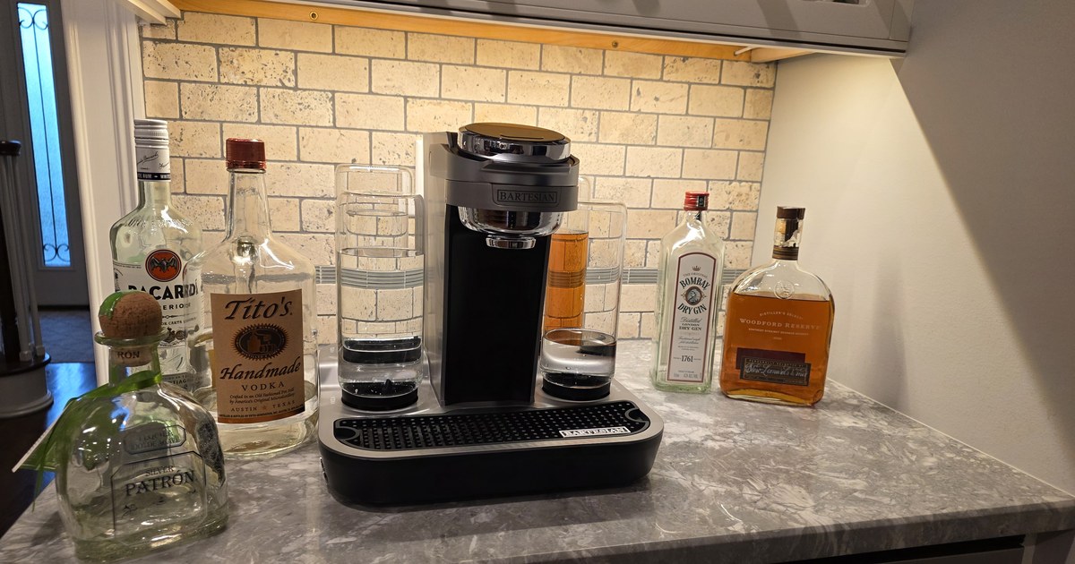 Bartesian cocktail machine on a granite countertop surrounded by Patrón, Bacardi, Tito's, Bombay Dry Gin, and Woodford Reserve — a hosting-ready home bar setup with stone backsplash and warm under-cabinet lighting.