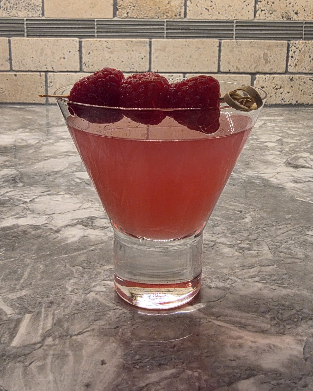 A coral Raspberry Ginger Gimlet gin cocktail garnished with three raspberries on a pick.