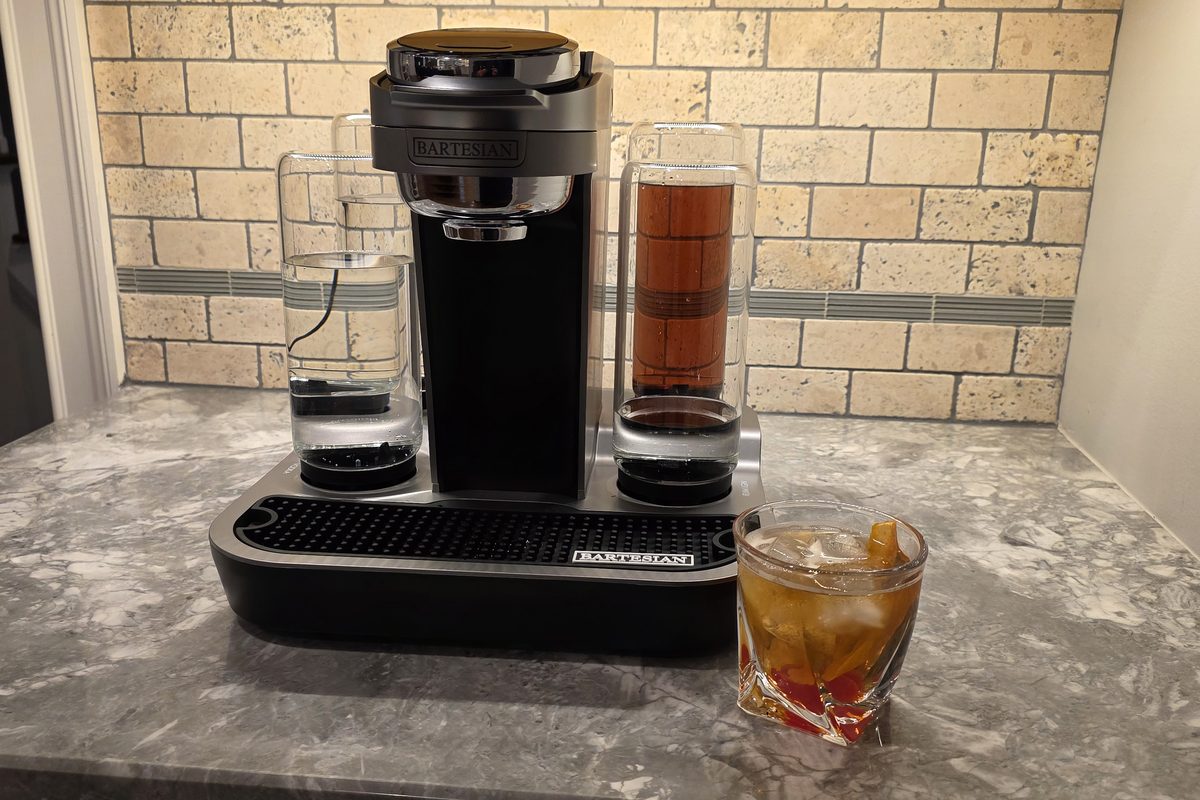 A finished Bartesian Old Fashioned over ice with an orange peel and cherries, next to the Bartesian machine on a marble counter