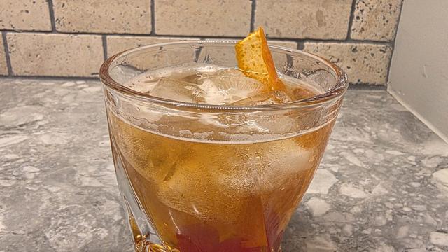 Bartesian Old Fashioned cocktail in a heavy rocks glass over ice, garnished with an orange peel and maraschino cherries on a marble counter