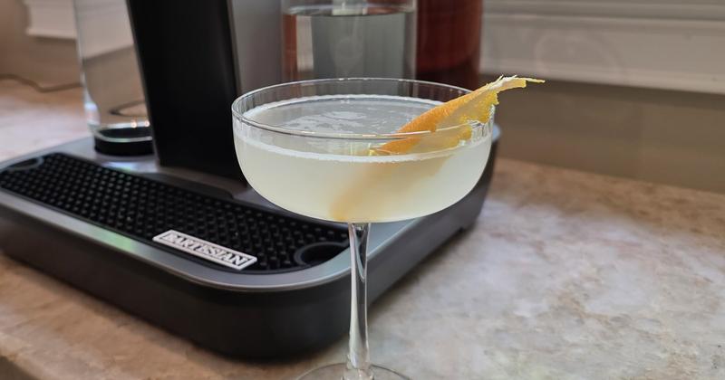A Bee's Knees in a coupe with a lemon peel, beside a Bartesian machine on a stone counter