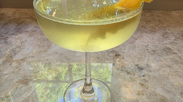 Bartesian Uptown Rocks cocktail in a coupe glass with a soft pale-yellow color and a lemon peel garnish on a marble counter by a sunlit window