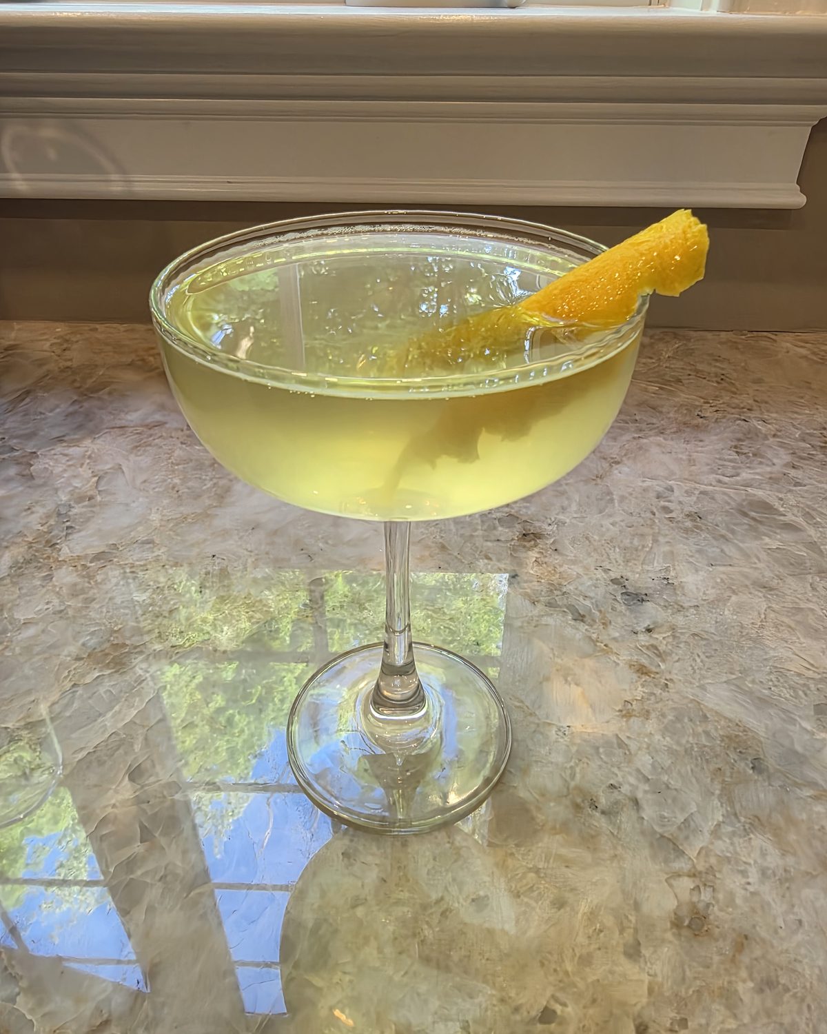 Bartesian Uptown Rocks cocktail in a coupe glass with a soft pale-yellow color and a lemon peel garnish, served on a marble counter by a sunlit window