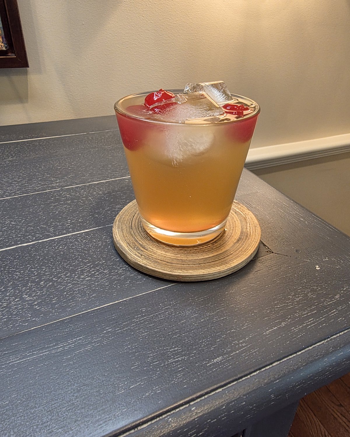 Bartesian Whiskey Sour cocktail in a low ball glass, hazy light amber with maraschino cherries and ice, on a wood coaster in a home bar corner